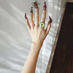 May include: A hand features long, glossy red fingernails adorned with silver-toned metal charms, including a spider and chains. A round ring with a pale green background and a black scorpion graphic is worn on the ring finger.