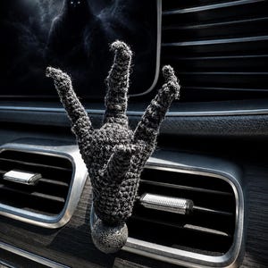 May include: A dark gray, three-pronged crochet claw ornament attached to a textured, spherical base. The object is mounted onto the horizontal slats of a vehicle air vent, positioned in front of a screen displaying a hooded figure under a full moon.