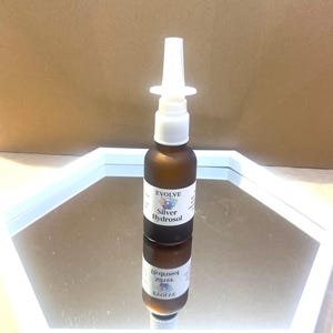 May include: A brown glass bottle with a white nasal spray applicator. The bottle has a label that reads "EVOLVE Silver Hydrosol". The bottle is reflected in a mirror. The background is a light brown color.