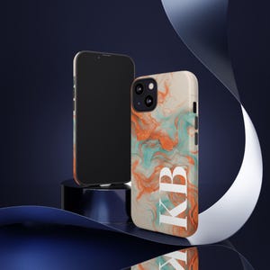 May include: A smartphone and a phone case with a colorful abstract design. The case features a cream background with orange and teal swirls and the white letters "KB". The phone is black.