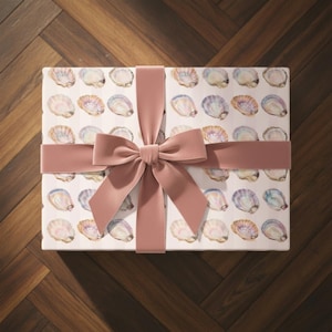 Pode incluir: Uma caixa de presente embrulhada com um padrão de conchas em aquarela em tons de azul, rosa e dourado. Uma fita e um laço de veludo rosa empoeirado completam a apresentação, criando um visual sofisticado.