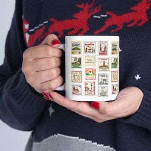 May include: White ceramic mug featuring a collage of vintage-style travel stamps. The stamps showcase various destinations, including Brooklyn, Canada, and France. The mug is held by a person wearing a navy blue sweater with a red and white reindeer pattern.