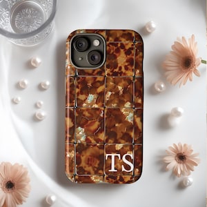 May include: A brown and gold phone case with a tortoiseshell pattern and the initials "TS" in white. The case has a square tile design. The image also includes pearls and pink flowers.