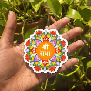 Puede incluir: Una colorida pegatina con un diseño floral y el texto hindi "Shri Radha" en el centro. La pegatina presenta una vibrante gama de colores, incluyendo naranja, rojo, amarillo, azul y morado. La pegatina se sostiene en una mano sobre un fondo de plantas verdes.