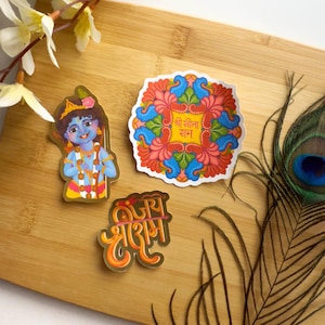 Puede incluir: Tres pegatinas coloridas sobre una superficie de madera. Una pegatina representa una deidad de piel azul, otra presenta un diseño floral con texto y la tercera tiene escritura naranja. También se ven una pluma de pavo real y detalles florales.