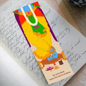 May include: A colorful bookmark featuring a cartoon illustration of a deity with blue skin, wearing yellow and green garments. The bookmark has the text "Do your best, leave the rest to krishna" at the bottom. The bookmark is placed on a book with handwritten text.