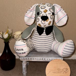 May include: A gray and white striped dog plushie with a black bow tie. The dog's head is made from a white fabric with a yellow and brown animal print. The dog's paws are pink and white striped. The dog is sitting on a white stool with a wooden top. The stool is sitting on a brown carpet. A wooden circle with the text "Sinais Beauty Handmade Clothes" is in the foreground.