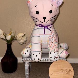 May include: A pink and white patchwork cat plush toy with a purple bow and black polka dots on its paws. The cat is sitting on a small white stool with a wooden round sign that says "Sinais Beauty Handmade Clothes" with a heart and the initials "SB".