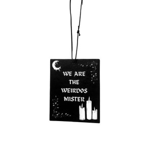 May include: Black car air freshener with white text that reads "We are the weirdos mister" and a crescent moon with dripping wax. Three white candles are on the right side of the air freshener.