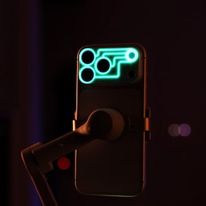 May include: A smartphone with a glowing green outline around the camera lenses. The phone is mounted on a gimbal stabilizer, with a dark background and bokeh lights.