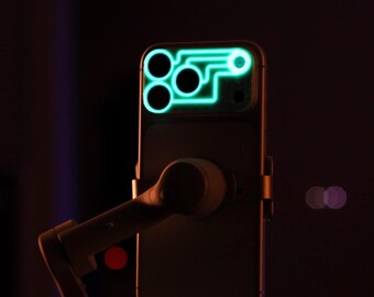 Könnte beinhalten: Ein Smartphone mit einer leuchtend grünen Umrandung um die Kameralinsen. Das Telefon ist an einem Kardan-Stabilisator befestigt, mit dunklem Hintergrund und Bokeh-Lichtern.