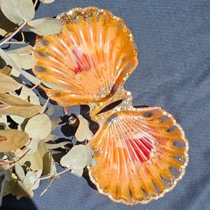 May include: Two orange and gold seashell-shaped resin dishes with red accents. The dishes are nested together.