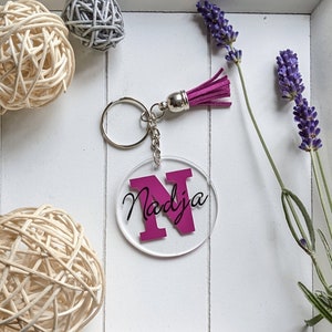 Puede incluir: Un llavero redondo de acrílico transparente con la letra "N" magenta y el nombre "Nadja" en escritura cursiva. Tiene una cadena y un anillo plateados, y una borla magenta. Elementos decorativos incluyen lavanda y esferas tejidas.