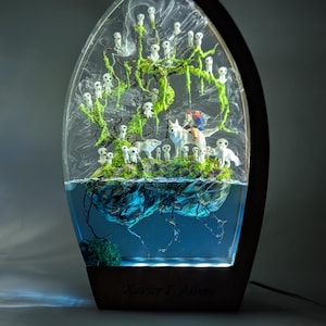 Könnte beinhalten: Dekorative Harzlampe mit einer Miniatur-Waldszene. Die Szene zeigt einen Baum mit moosbewachsenen Ästen, weiße Geisterfiguren und eine Figur, die auf einem Wolf reitet. Der Sockel ist aus Holz, und die Lampe hat einen blauen Wassereffekt.