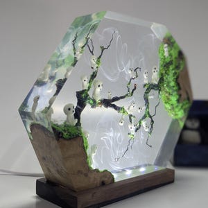 Pu&ograve; includere: Una lampada esagonale in resina con una base in legno. La resina contiene una scena di foresta in miniatura con un albero, muschio e piccole figure bianche. La lampada &egrave; accesa e diffonde una luce calda.