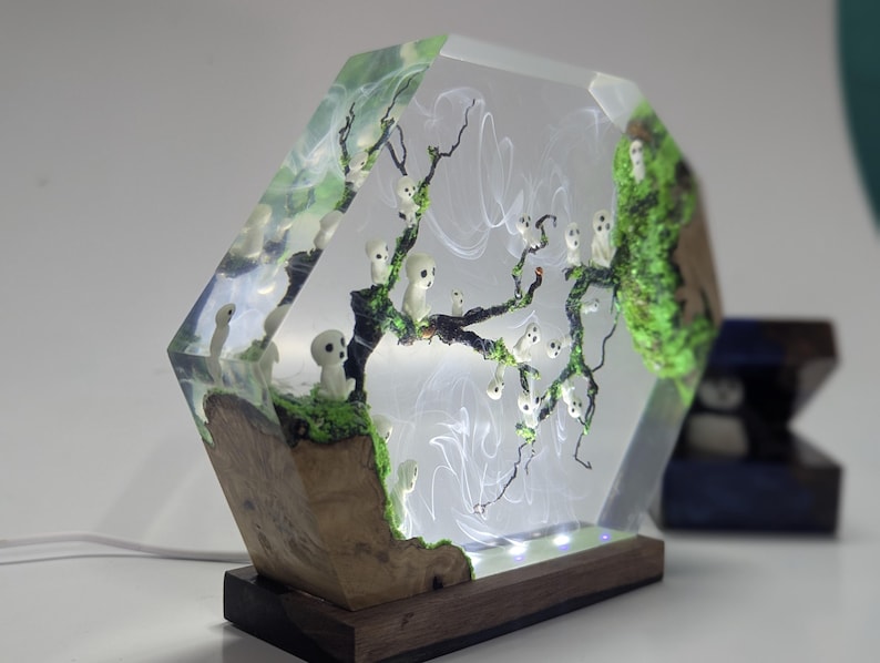 Pu&ograve; includere: Una lampada esagonale in resina con una base in legno. La resina contiene una scena di foresta in miniatura con un albero, muschio e piccole figure bianche. La lampada &egrave; accesa e diffonde una luce calda.