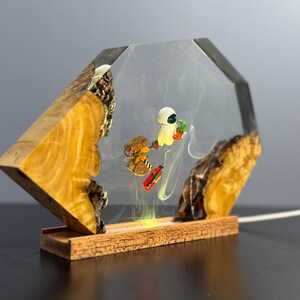 May include: A decorative lamp featuring a clear resin enclosure with a wooden base and accents. Inside, miniature figures of a robot and a character holding a plant are suspended. The lamp has a warm, yellow glow.