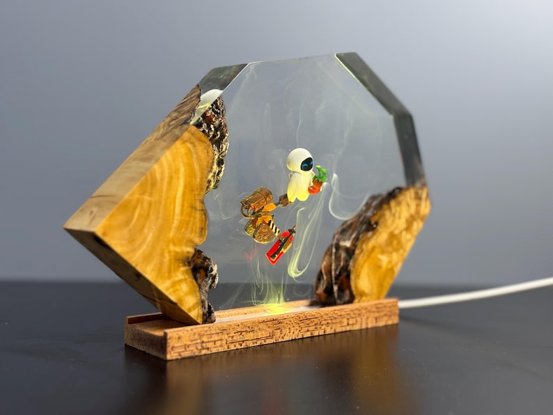 May include: A decorative lamp featuring a clear resin enclosure with a wooden base and accents. Inside, miniature figures of a robot and a character holding a plant are suspended. The lamp has a warm, yellow glow.
