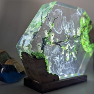 Pu&ograve; includere: Una lampada in resina esagonale con una base trasparente e un interno verde e bianco. L'interno presenta un albero con figure bianche e vortici bianchi. La lampada &egrave; accesa e ha una base in legno.