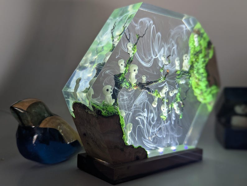 Pu&ograve; includere: Una lampada in resina esagonale con una base trasparente e un interno verde e bianco. L'interno presenta un albero con figure bianche e vortici bianchi. La lampada &egrave; accesa e ha una base in legno.