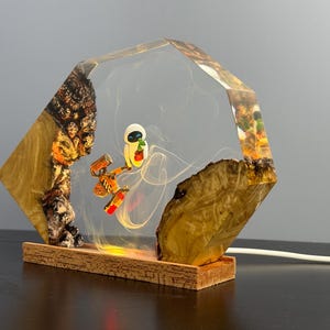 May include: A unique lamp featuring a clear, geometric resin encasing a cartoon character and a robot. The lamp has a wooden base and natural wood accents on the sides. The light glows from within, creating a warm ambiance.