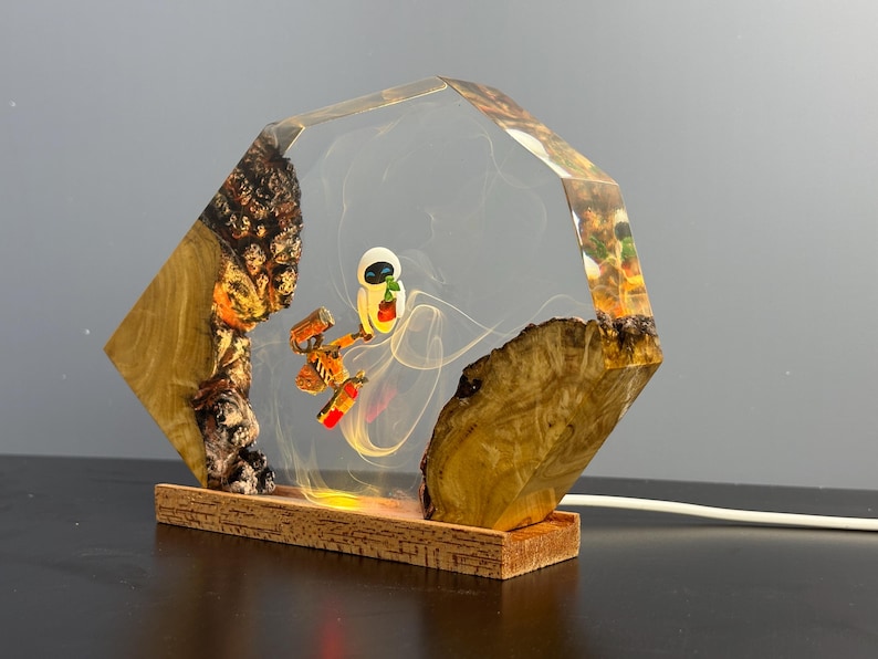 May include: A unique lamp featuring a clear, geometric resin encasing a cartoon character and a robot. The lamp has a wooden base and natural wood accents on the sides. The light glows from within, creating a warm ambiance.