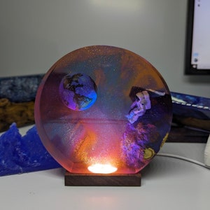 Op de afbeelding: Een ronde harslamp met een galaxy-ontwerp. De lamp heeft een kleine globe en een astronautbeeldje. De lamp wordt van onderen verlicht en geeft een warm gloed.
