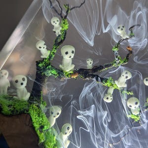 Pu&ograve; includere: Un'opera d'arte in resina con un albero con figure bianche luminose e muschio verde. Le figure sono disposte lungo i rami dell'albero e sembrano fluttuare nell'aria.