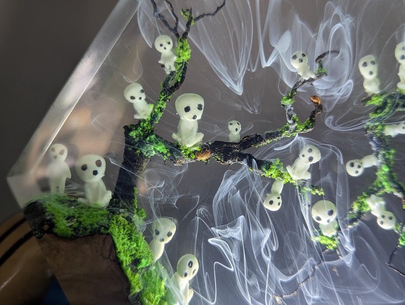 Pu&ograve; includere: Un'opera d'arte in resina con un albero con figure bianche luminose e muschio verde. Le figure sono disposte lungo i rami dell'albero e sembrano fluttuare nell'aria.
