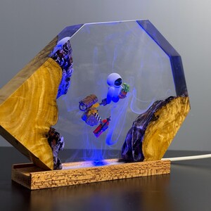 May include: A unique lamp featuring a wooden base and a clear, geometric resin structure. Inside, a blue-lit scene depicts a robot with a plant. The wooden elements have a natural, warm tone, contrasting with the cool blue light and resin.