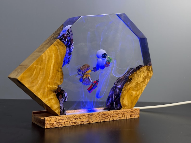May include: A unique lamp featuring a wooden base and a clear, geometric resin structure. Inside, a blue-lit scene depicts a robot with a plant. The wooden elements have a natural, warm tone, contrasting with the cool blue light and resin.