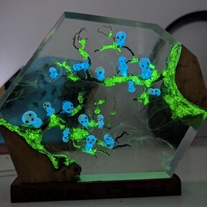 Pu&ograve; includere: Una scultura esagonale in resina con un albero verde luminoso e figure blu luminose. La scultura &egrave; su una base di legno.