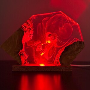 May include: A unique, geometric-shaped lamp with a red glow. The lamp features a clear, resin-like center with an abstract design, framed by natural wood elements. The base is a rectangular wooden platform.