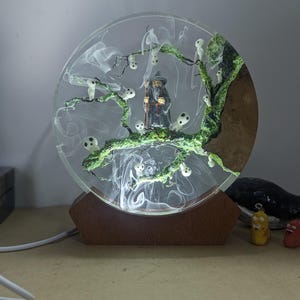 以下が含まれることがあります： 円形に照らされた樹脂アート作品。ミニチュアの魔法使いの姿が、光る幽霊のような姿の木の下に立っています。木は緑色の苔で詳細に描かれ、樹脂は煙のような効果があります。ベースは木製です。