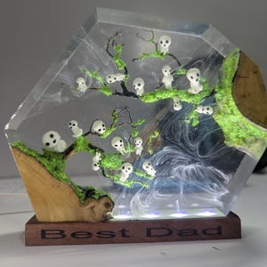 Pu&ograve; includere: Una scultura in resina trasparente di un albero con figure bianche e muschio verde. La base &egrave; in legno e ha le parole "Best Dad" incise.