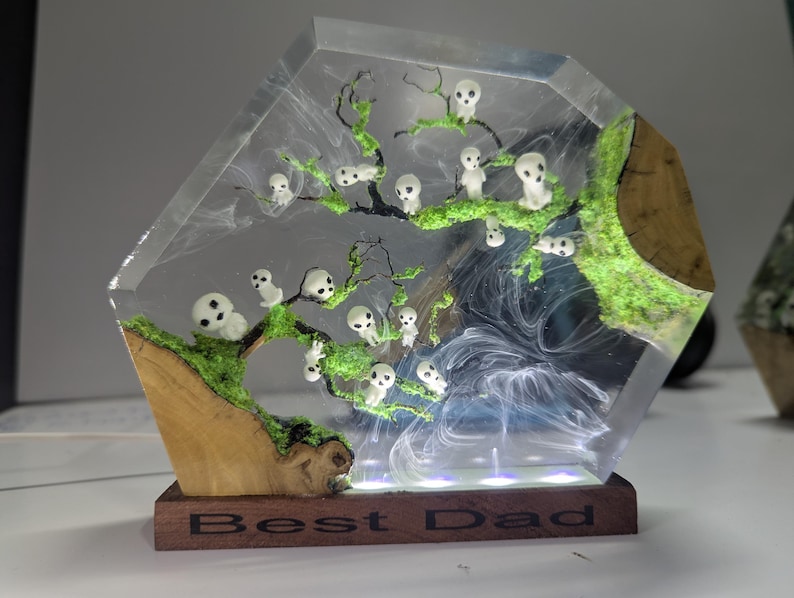Pu&ograve; includere: Una scultura in resina trasparente di un albero con figure bianche e muschio verde. La base &egrave; in legno e ha le parole "Best Dad" incise.