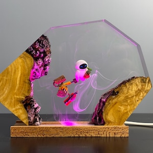 May include: A decorative lamp featuring a clear, geometric resin design with a wooden base. Inside, a small robot figure is illuminated with pink light, set against a backdrop of swirling pink smoke. The lamp's wooden elements have a natural, textured appearance.