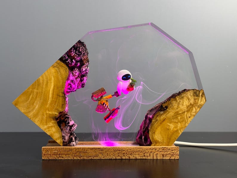 May include: A decorative lamp featuring a clear, geometric resin design with a wooden base. Inside, a small robot figure is illuminated with pink light, set against a backdrop of swirling pink smoke. The lamp's wooden elements have a natural, textured appearance.