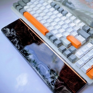 Peut inclure: Un clavier mécanique blanc et gris avec des touches d'accent orange et un repose-poignet assorti. Le repose-poignet présente un motif sombre et tourbillonnant. Le clavier a un design minimaliste avec une disposition compacte.
