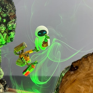 May include: A whimsical artwork featuring the animated characters Wall-E and Eve. Eve is depicted in a glowing green hue, holding a small object. Wall-E is shown with a yellow and orange body, holding a bottle. The background has green swirls and wooden accents.