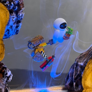 May include: A whimsical art piece featuring the characters Wall-E and Eve from the animated film. Eve, a white robot, holds a small plant. Wall-E, a small robot, is also present. The artwork is set against a smoky, blue-lit background, framed by wood.
