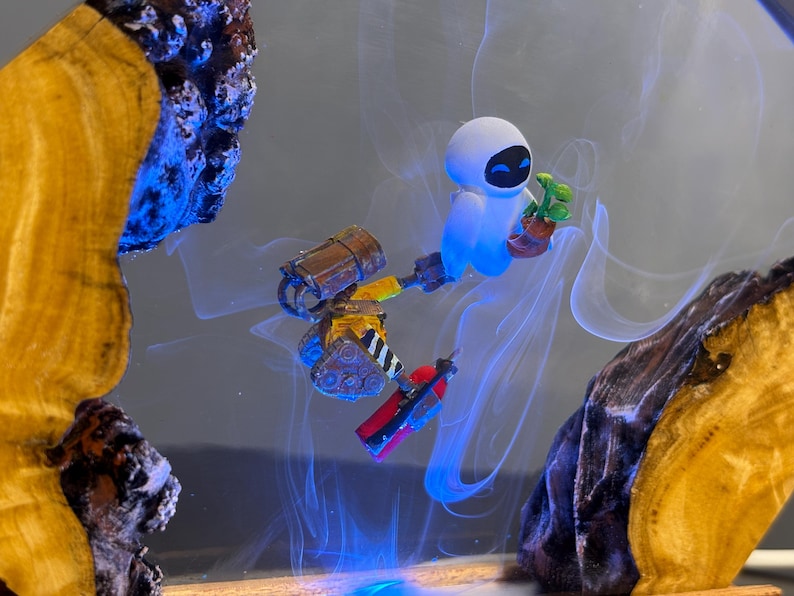 May include: A whimsical art piece featuring the characters Wall-E and Eve from the animated film. Eve, a white robot, holds a small plant. Wall-E, a small robot, is also present. The artwork is set against a smoky, blue-lit background, framed by wood.