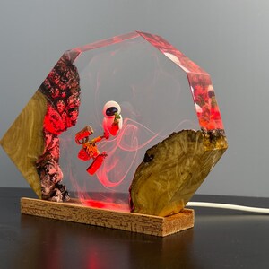 May include: A unique lamp featuring a clear, geometric resin design with a red and white character inside. The lamp is set on a wooden base and has a warm, red glow. The sides of the lamp are made of wood.