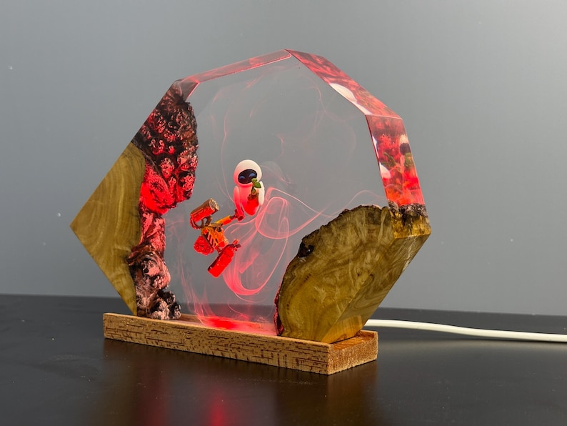 May include: A unique lamp featuring a clear, geometric resin design with a red and white character inside. The lamp is set on a wooden base and has a warm, red glow. The sides of the lamp are made of wood.