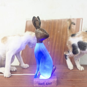 Puede incluir: Una escultura de conejo de madera con cabeza y orejas de color marrón oscuro y cuerpo azul brillante. La escultura se asienta sobre una pequeña base de madera con el texto "Wefi ART". La escultura está iluminada con luz azul.