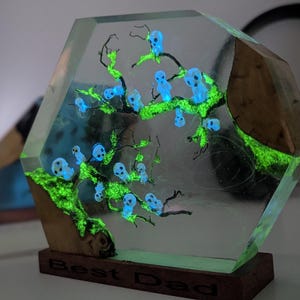 Pu&ograve; includere: Un blocco esagonale in resina trasparente con una base in legno. Il blocco contiene un albero verde fluorescente con figure blu fluorescenti. La base &egrave; incisa con le parole "Best Dad".