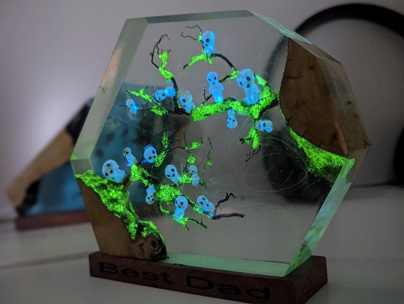 Pu&ograve; includere: Un blocco esagonale in resina trasparente con una base in legno. Il blocco contiene un albero verde fluorescente con figure blu fluorescenti. La base &egrave; incisa con le parole "Best Dad".
