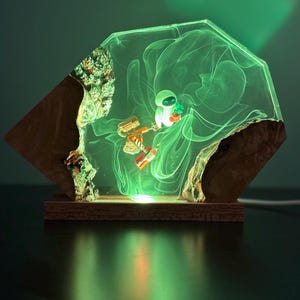 May include: A unique, illuminated resin lamp featuring a geometric design. The lamp showcases a small astronaut figure and a robot within a glowing green resin. The base is made of wood, and the lamp emits a soft, ambient light.