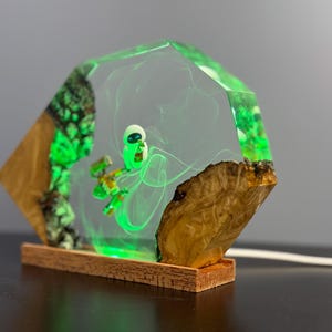 May include: A unique, handmade lamp featuring a green alien figure encased in clear resin. The lamp has a wooden base and sides with green accents, creating a glowing effect. The design incorporates natural wood elements.