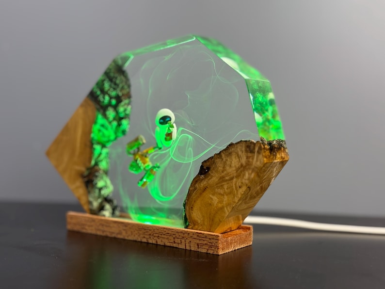 May include: A unique, handmade lamp featuring a green alien figure encased in clear resin. The lamp has a wooden base and sides with green accents, creating a glowing effect. The design incorporates natural wood elements.